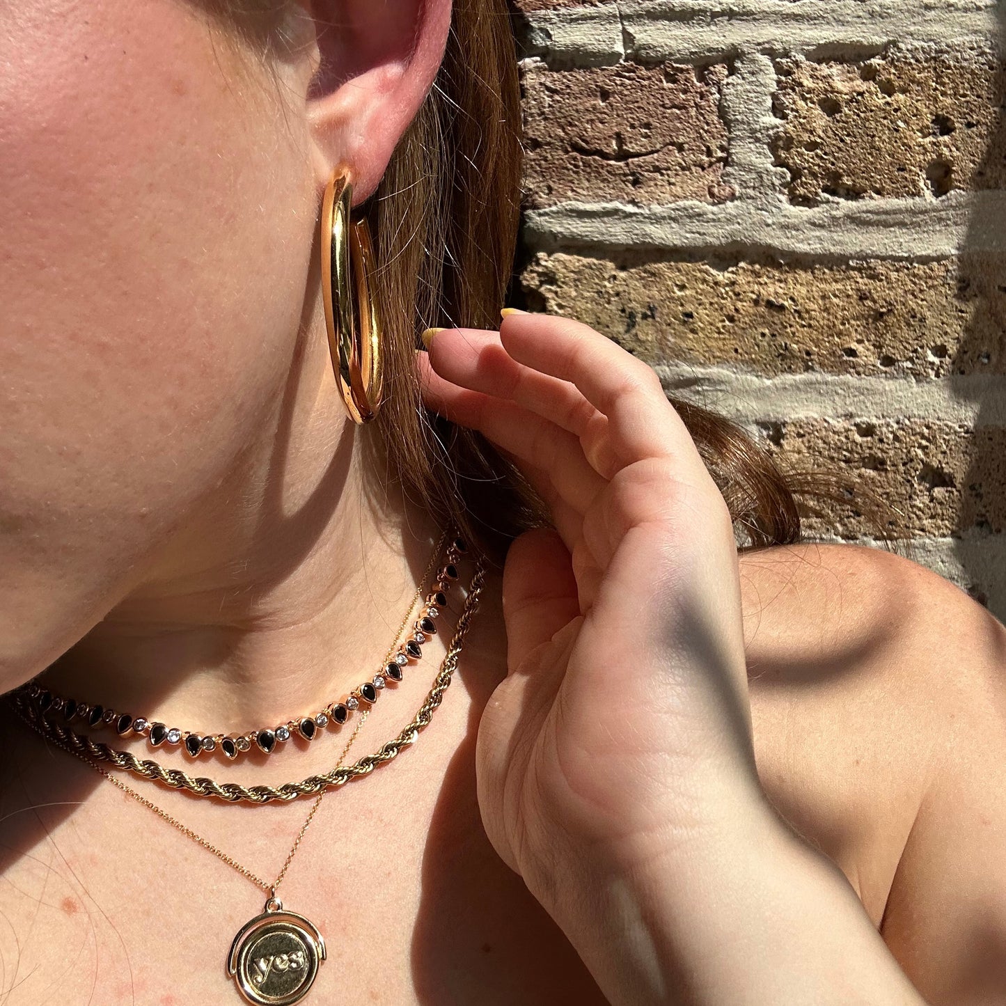 A person wearing large gold hoop earrings, a Teardrop Tennis Necklace, and a black beaded choker gently touches their ear as sunlight casts shadows on their skin with a textured brick wall in the background.