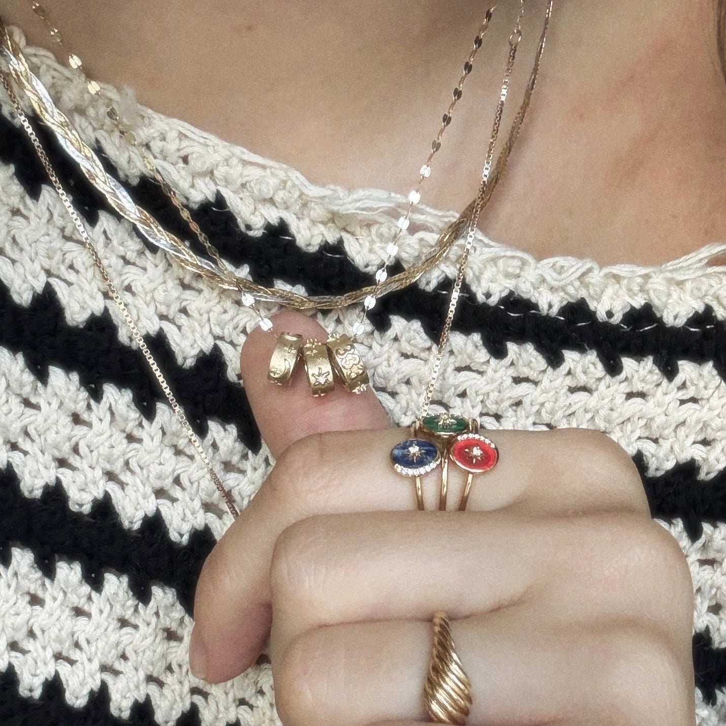 A person in a cream and black striped knit sweater displays several gold necklaces, the Solid Gold Angel Number Spacer Charm, gold rings, and three gemstone rings—blue, green, and red—on their fingers.