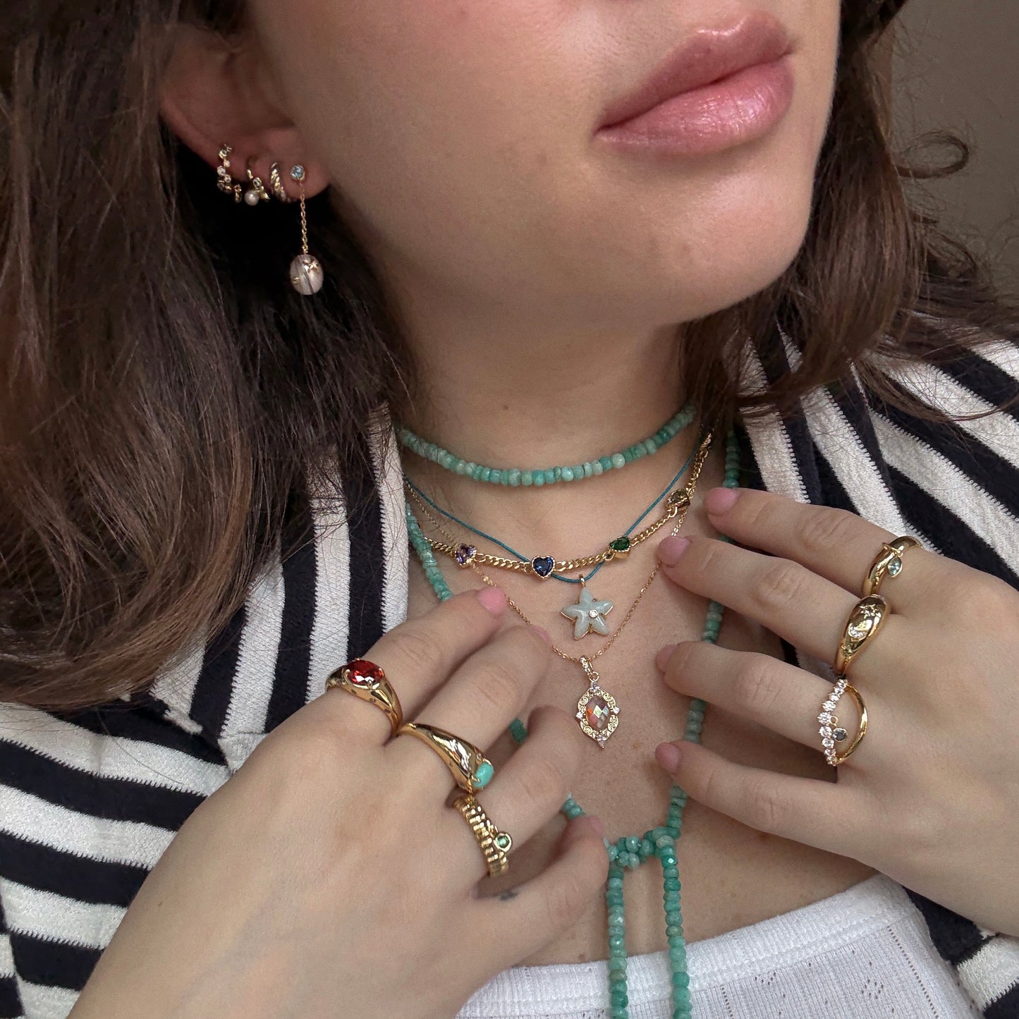 A woman showcases the Bold Fluted Charm Ring, layered beaded turquoise necklaces, two pendants, and earrings. Dressed in a black-and-white striped blazer, the focus is on her jewelry and lower face.