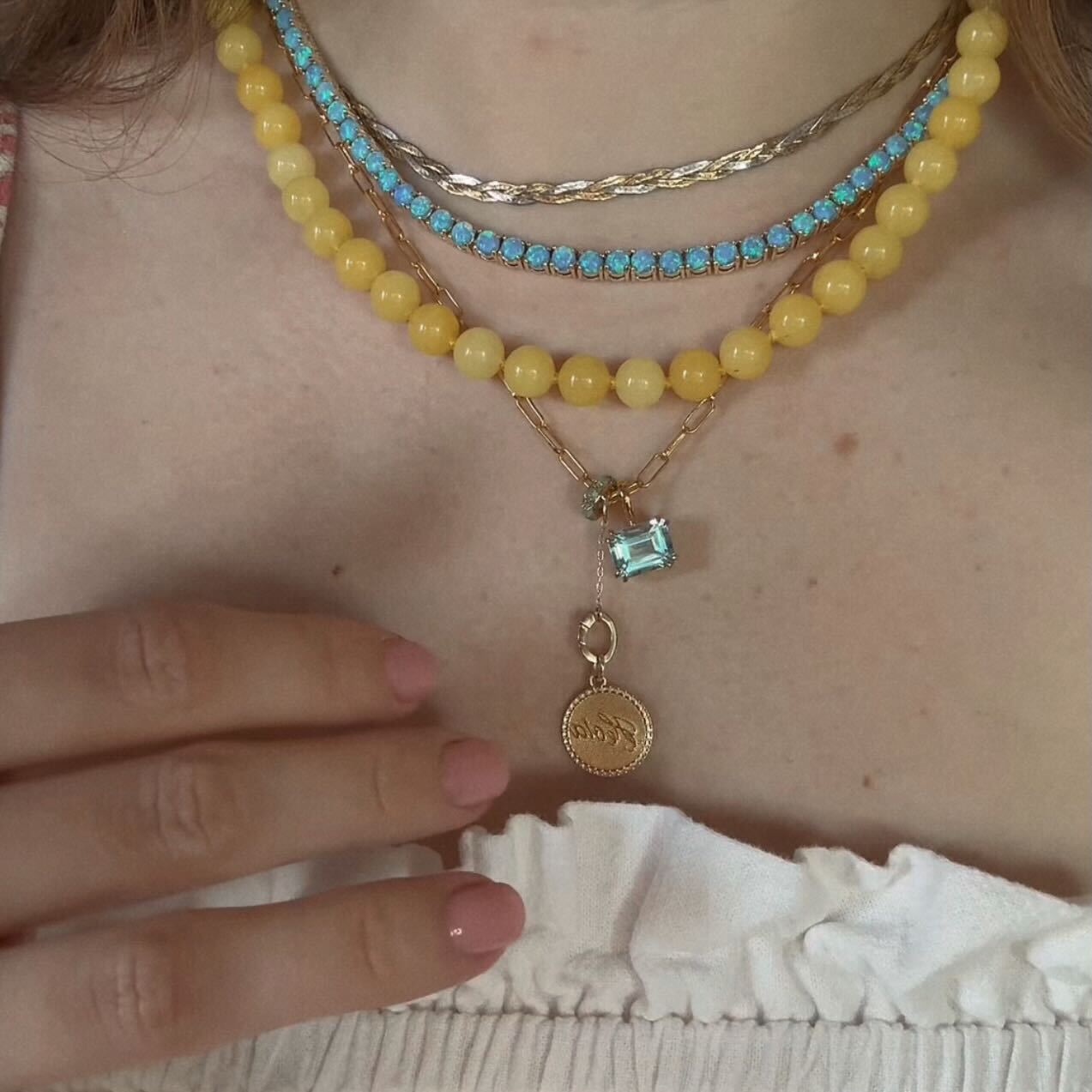 Wearing the Sale! Beaded Gemstone Necklace featuring yellow beads, turquoise stones, an 18k gold plated coin pendant, and a blue gemstone charm, their pink-polished hand rests on a white ruffled top.