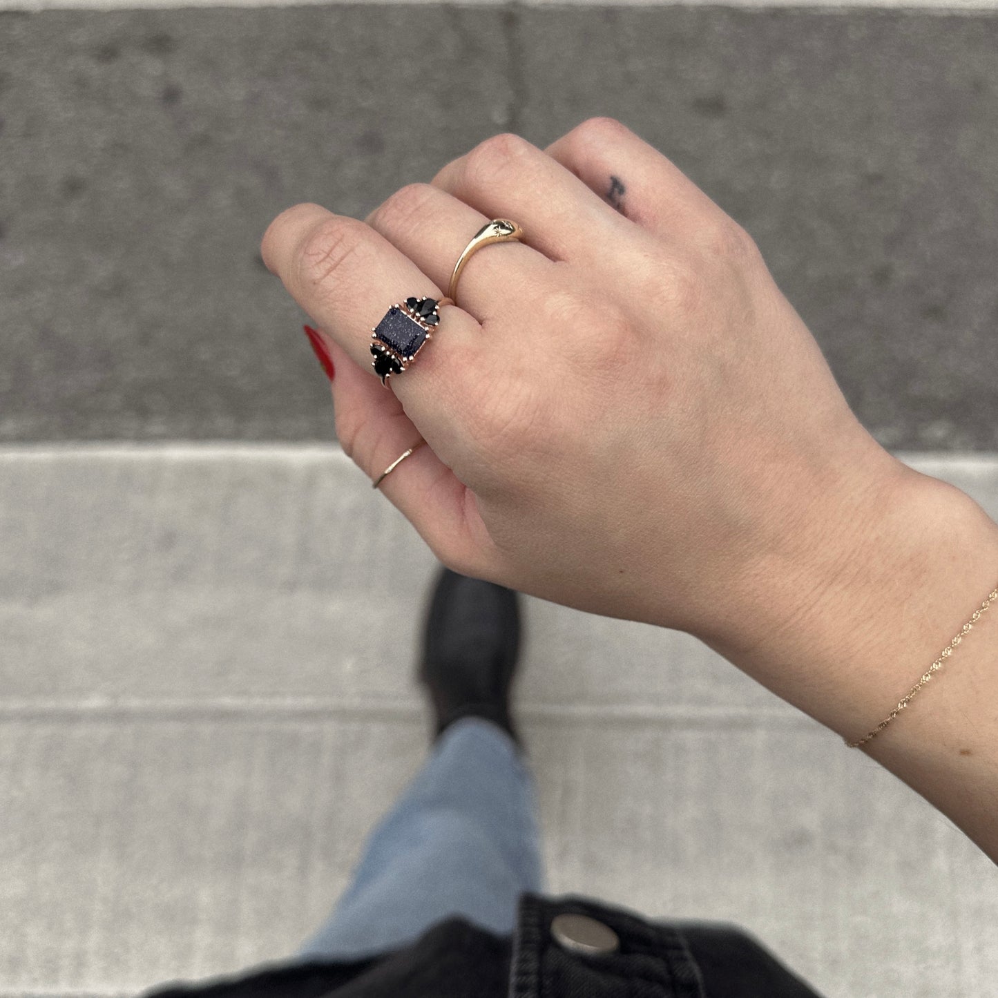 Wearing red nail polish and several rings, including the Gemstone Empress Ring with a striking black stone, this person also sports a delicate gold bracelet. A street and crosswalk are visible in the background.