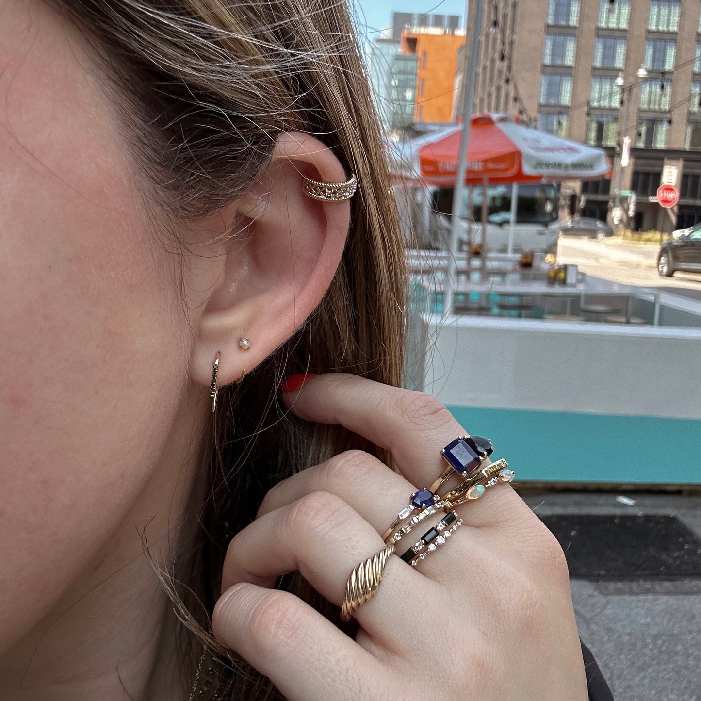 A close-up of a womans ear wearing Solid Gold Pearl Ear Huggers, her hand with gold and blue gemstone rings gently touching her hair. Urban buildings blur in the background, highlighting these earrings as perfect for everyday wear.