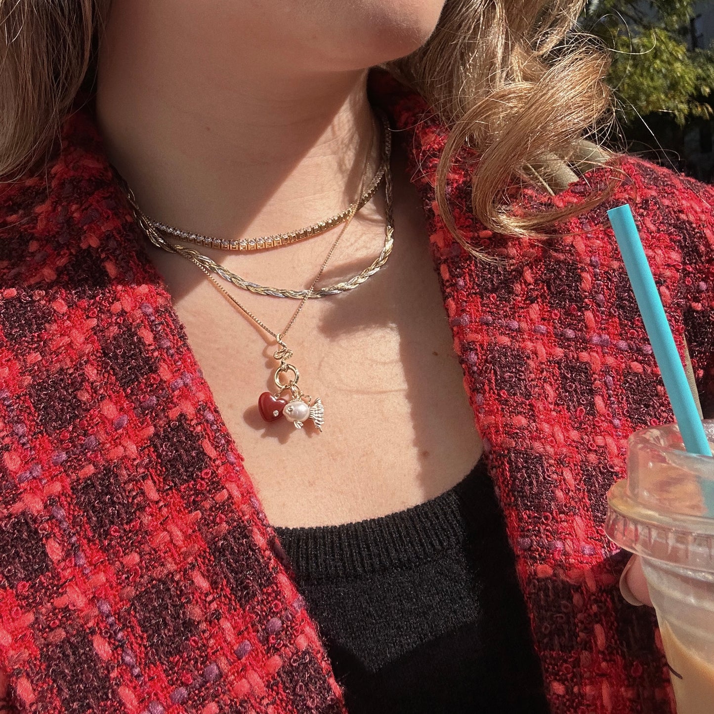 Wearing layered Solid Gold Statement Charms, including one with red and white accents, a person pairs them with a red textured jacket and holds a plastic cup with a blue straw; only their lower face and upper body appear in bright sunlight.