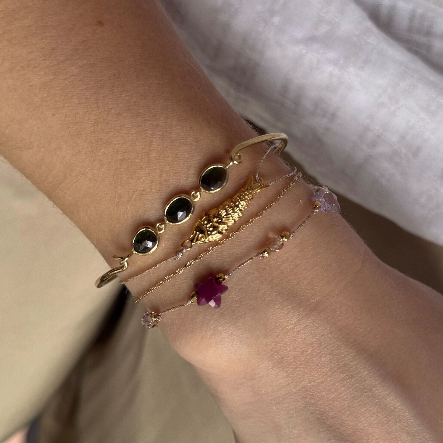 A wrist styled with three Sophie Deschamps Bijoux gold bracelets: one with black stones, one with a gold centerpiece, and the Sardina Bracelet—golden beads and a pink gemstone charm. The wearer has on a white top.