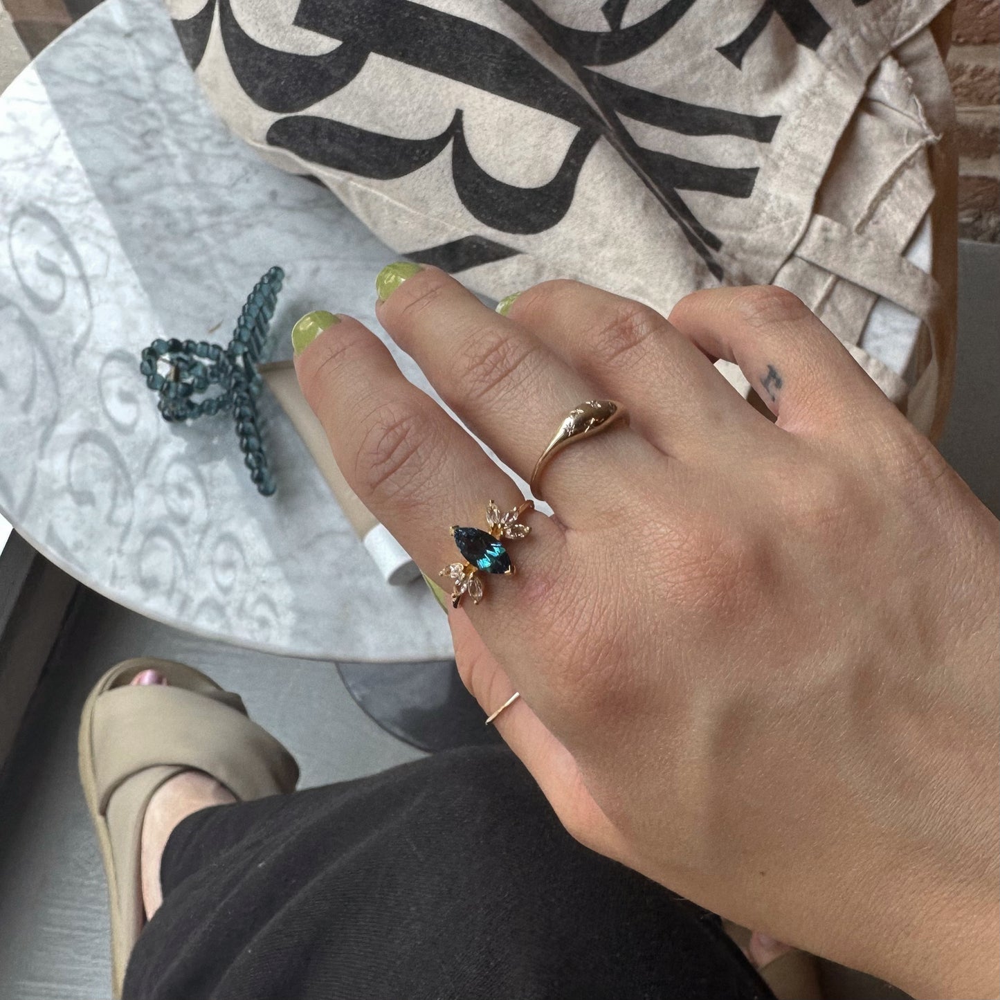 A hand with light green nail polish wears three yellow gold vermeil rings—including the Gemstone Firefly Ring—above a white marble table with a beaded bracelet and cloth bag. A foot in a beige sandal appears below.