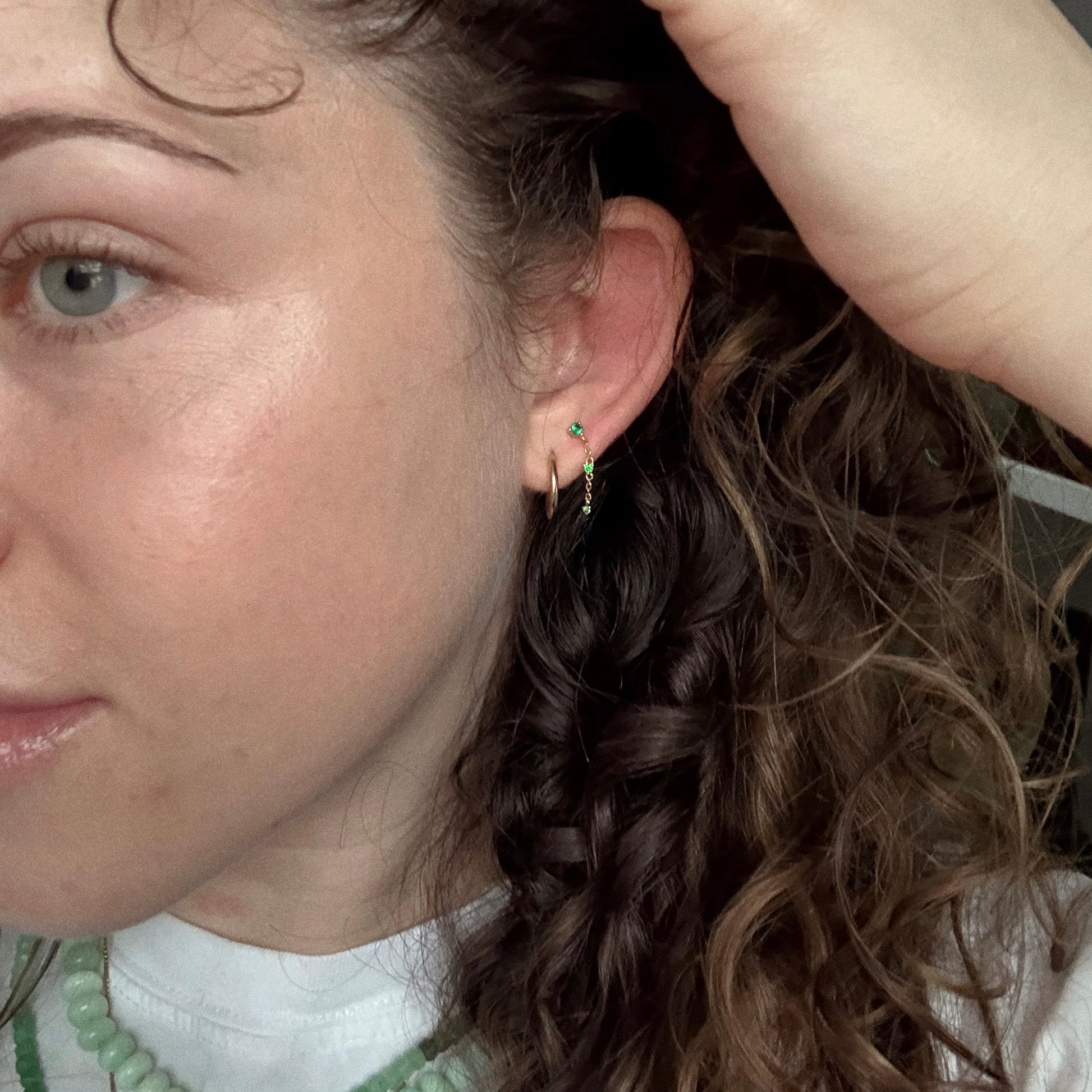 Close-up of a person with curly brown hair wearing Celebration Ombre Birthstone Drop Studs and green beaded necklaces over a white shirt, holding their hair back to reveal the sparkling earrings and part of their face.