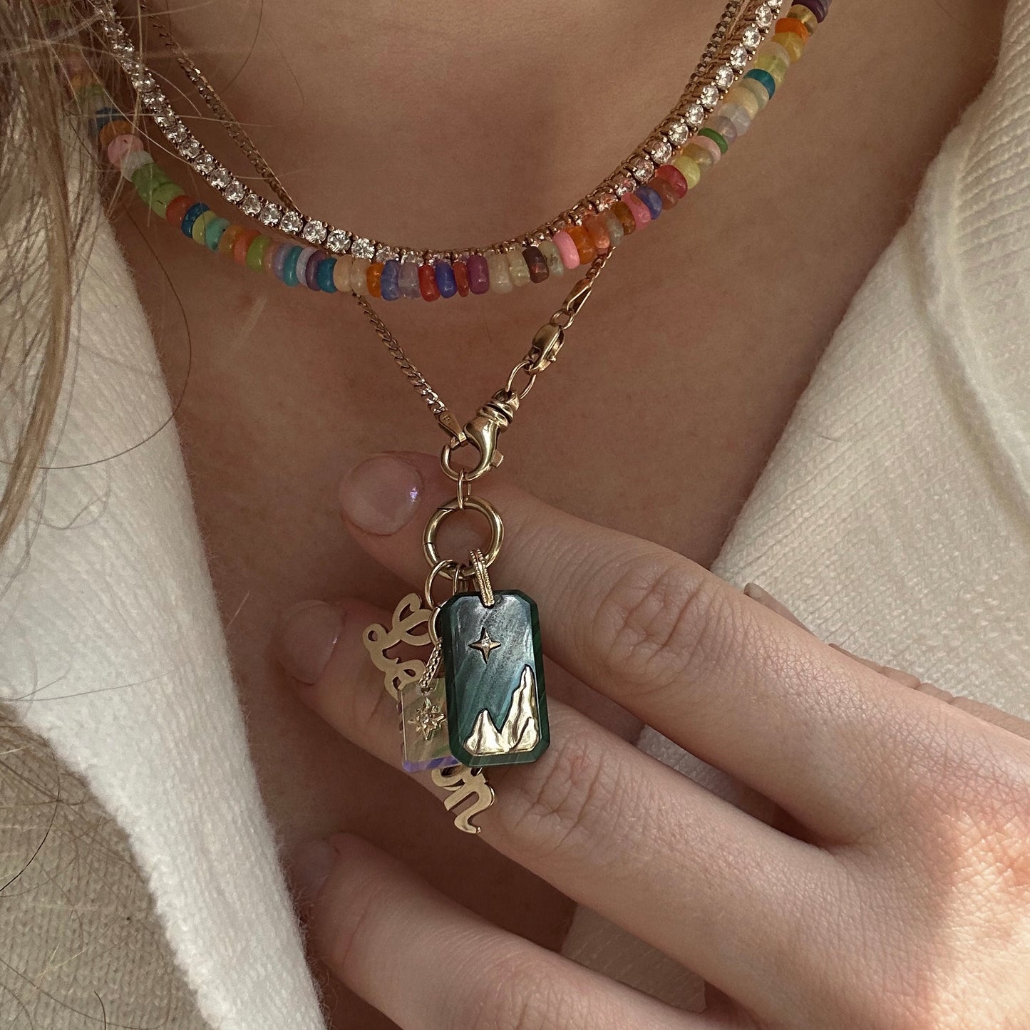 A close-up of a person wearing beaded and gold necklaces, including a mountain landscape pendant, a nameplate necklace, and the Solid Gold Crystal Element Charm. The pendants are shown against a cream-colored textured top.