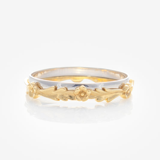 The Solid Gold Mixed Metal Filigree Band features a slim, two-toned gold band with a silver base and intricate gold floral filigree, showcasing small leaves and a central flower, set against a plain white background.