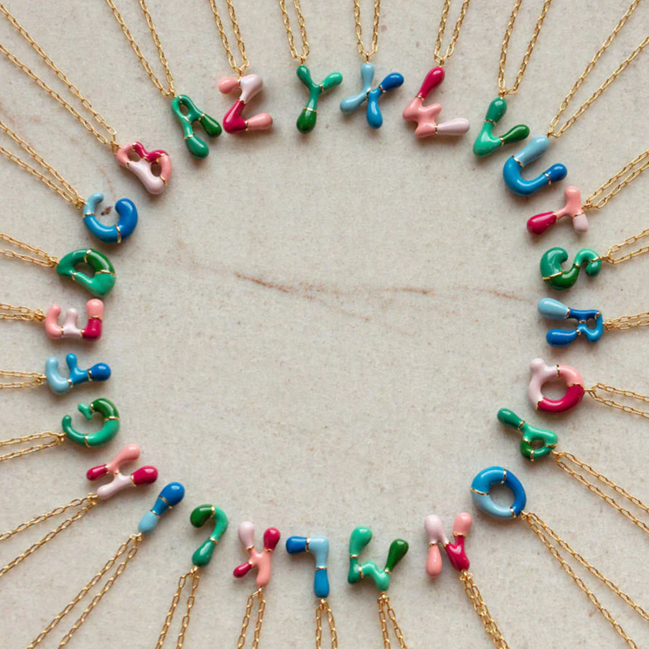 The Bubble Letter Necklace features playful bubble letter pendants in blue, pink, green, or white enamel on a 14k gold-plated chain, beautifully arranged in a circle.