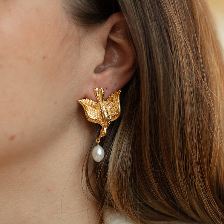 A close-up of a womans ear wearing Sparrow Earrings in gold vermeil, featuring detailed wings and a dangling white pearl. Her brown hair partially covers her ear, highlighting the elegance of these unique earrings.
