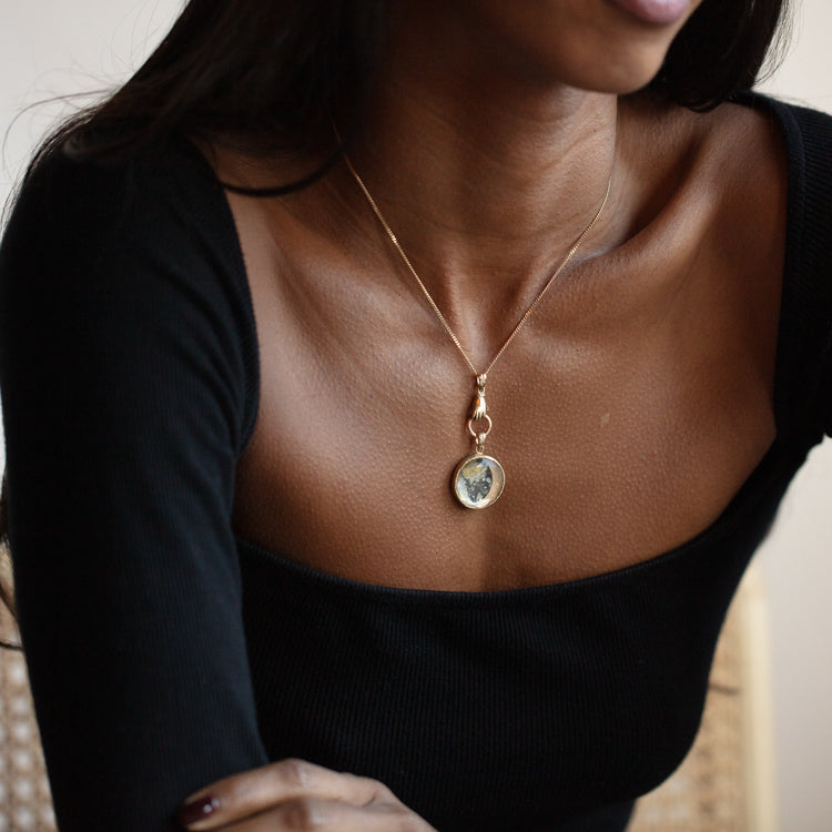 A woman sits with arms folded, her collarbone and part of her face in soft light, wearing a black square-neck top and the 14k Solid Gold Tiny Charms necklace by Acanthus.