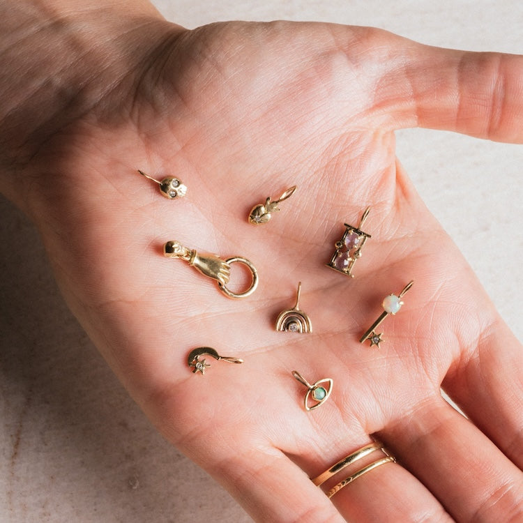A hand with two gold rings shows eight 14k solid gold tiny charms by Acanthus—featuring stars, a crescent moon, a planet, and a gemstone—in the palm.