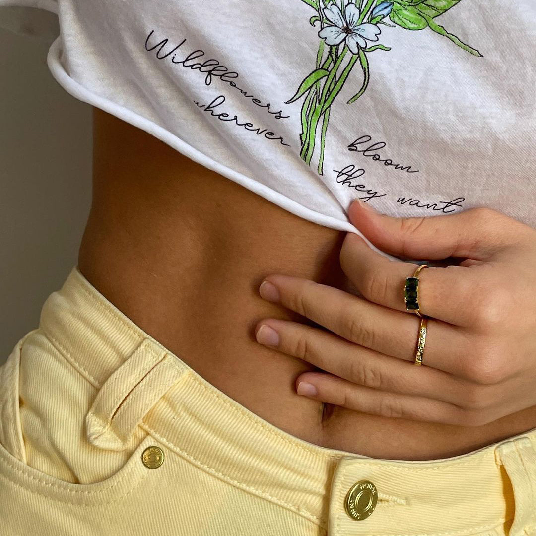 A person in yellow pants and a floral white shirt reading Wildflowers bloom wherever they want lifts their shirt to reveal their midriff and wears the Simple Star Set Diamond Band on their pinky finger.