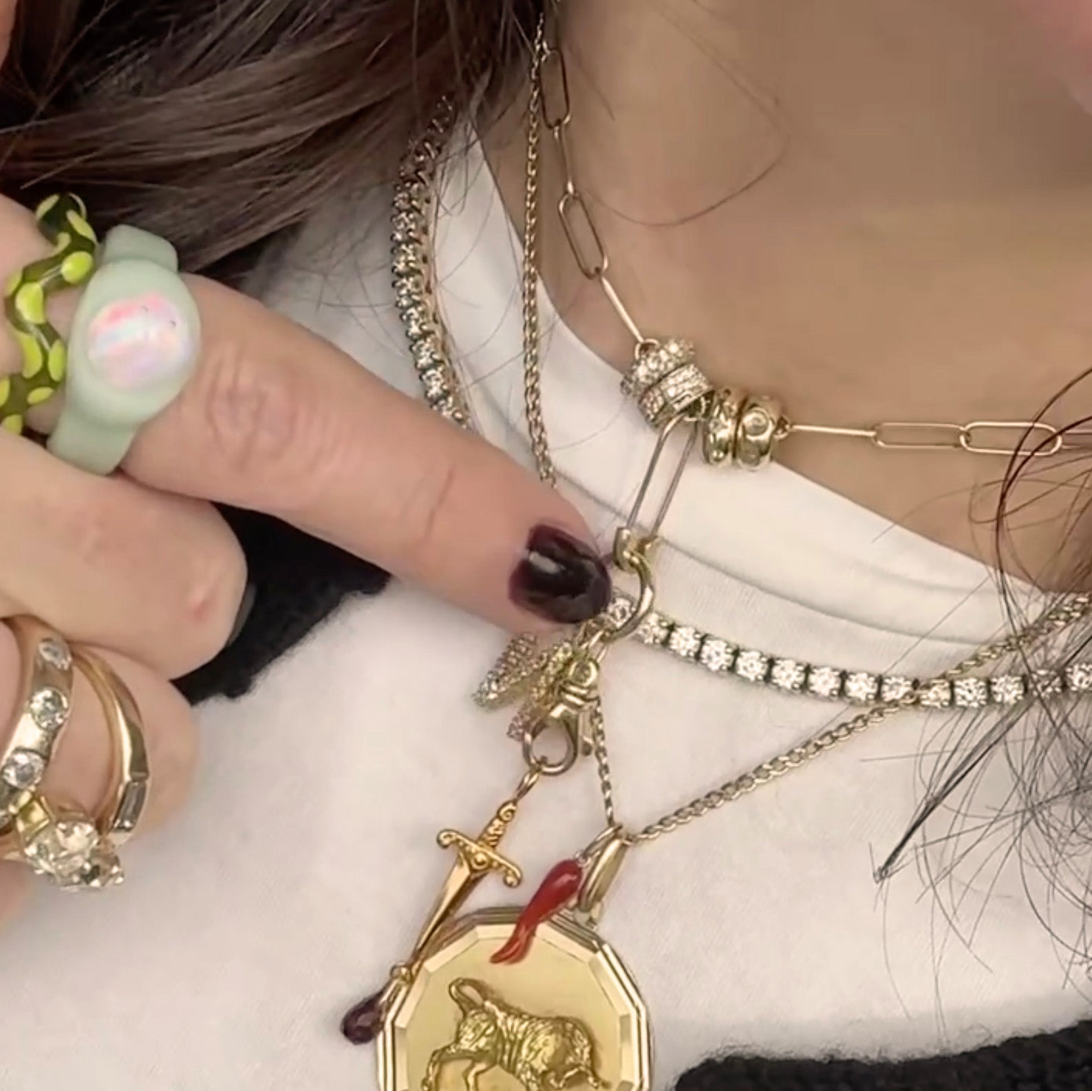 A close-up of someone wearing layered gold and diamond necklaces, chunky rings, and a light green ring with a pink stone. They point to a pendant with a lion and sword, accented by yellow gold charms and the Solid Gold Spacer Charm.