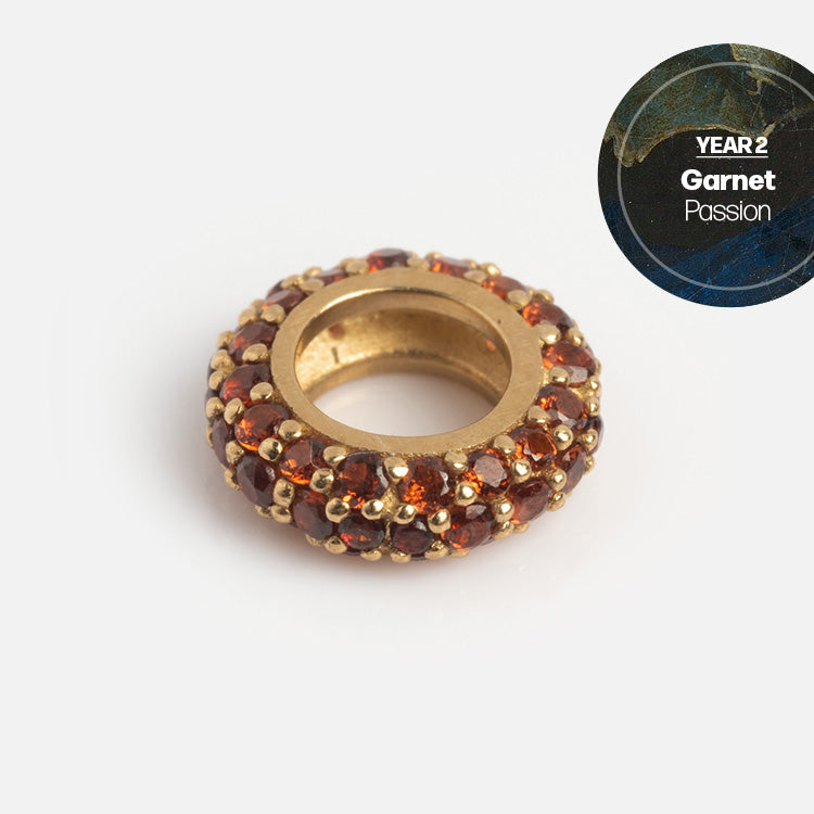 A gold, ring-shaped bead adorned with small garnet gemstones is displayed on a white background. In the top right, a circular label reads “YEAR 2 Garnet Passion.” Discover our Solid Gold Anniversaire Charm—ideal milestone jewelry.