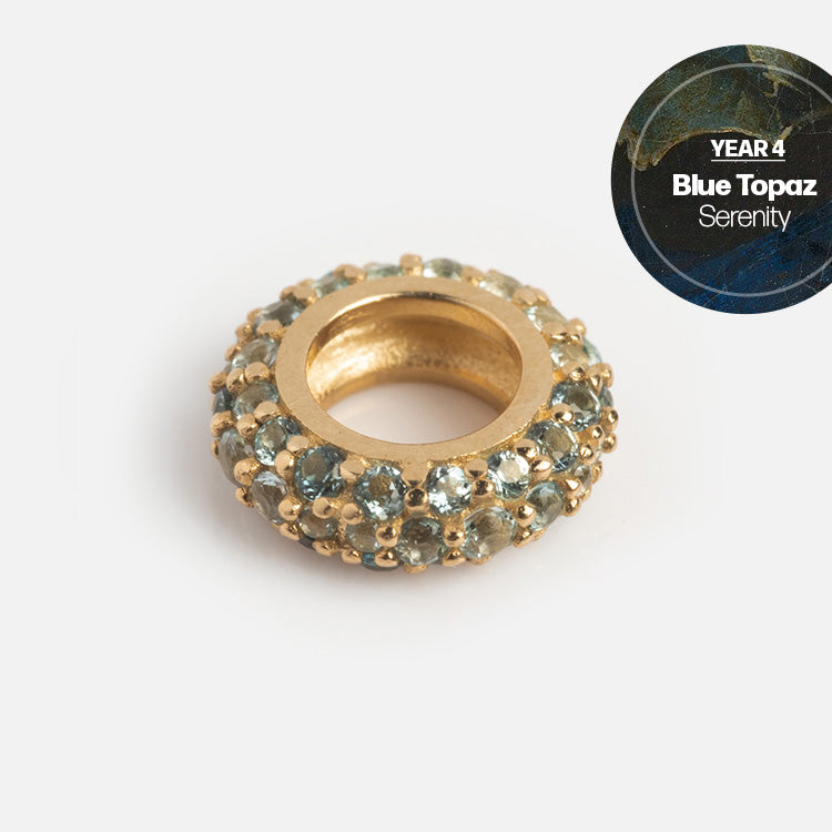 The Solid Gold Anniversaire Charm is a round gold piece with blue topaz gemstones, shown on a white background. A circular label in the top right reads YEAR 4 Blue Topaz Serenity—an ideal milestone jewelry gift.