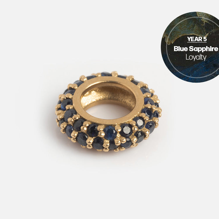 The Solid Gold Anniversaire Charm, a circular design adorned with blue sapphires, shines against a light backdrop. In the top right, “YEAR 5 Blue Sapphire Loyalty” appears on a marbled blue label—ideal milestone jewelry.