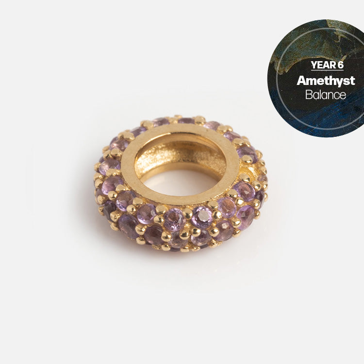 A Solid Gold Anniversaire Charm: a circular gold bead set with small purple amethyst stones. In the top right, a round label says YEAR 6 Amethyst Balance against a dark marble-like background—a beautiful milestone jewelry piece.