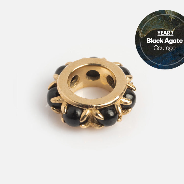 The Solid Gold Anniversaire Charm features black agate stones set in a gold circular frame with decorative detail and a YEAR 7 Black Agate Courage badge, making it the perfect milestone jewelry piece.