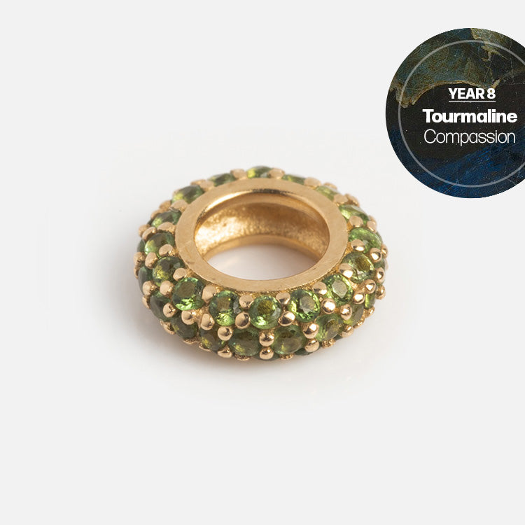 A Solid Gold Anniversaire Charm, round and bead-shaped, set with small green tourmalines. A circle in the corner reads YEAR 8 Tourmaline Compassion on a blue and gold marbled background—ideal for milestone or anniversary jewelry.