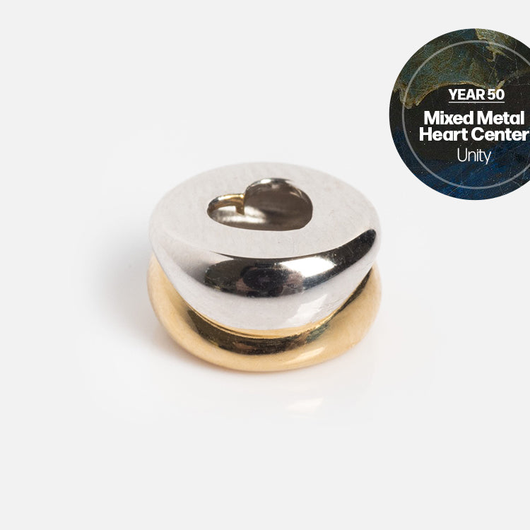 A small, round mixed-metal bead with a silver top featuring a heart cutout and a gold bottom. Labeled “Solid Gold Anniversaire Charm,” this milestone jewelry piece displays “Unity” on a circular badge in the corner.