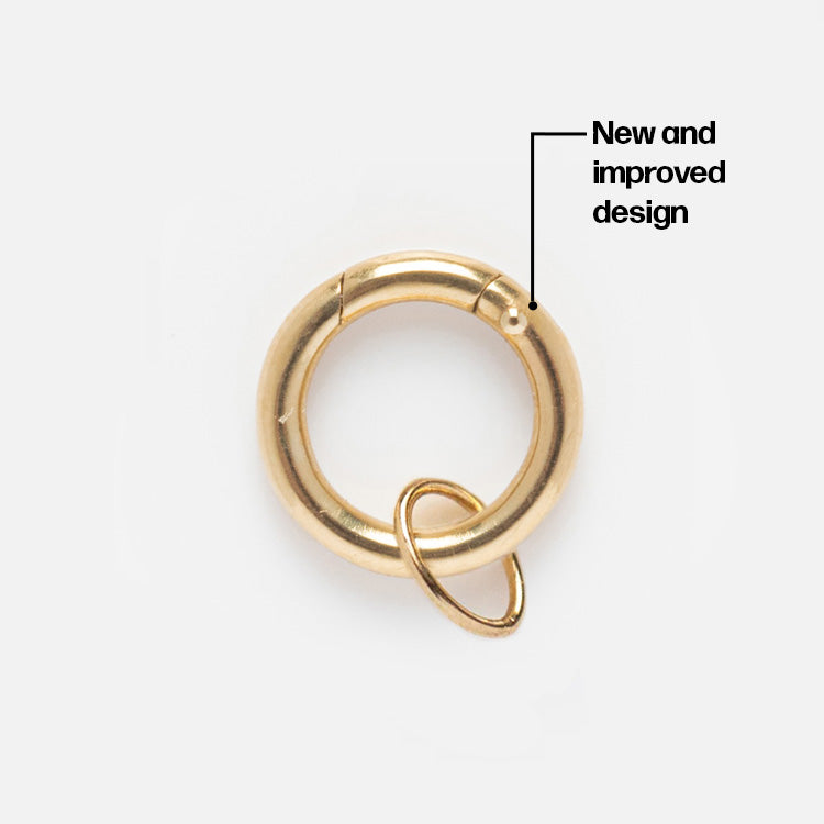 The Solid Gold Charm Clip features a circular clasp with a smaller ring for easy bracelet attachment, highlighted by an arrow labeled New and improved design.