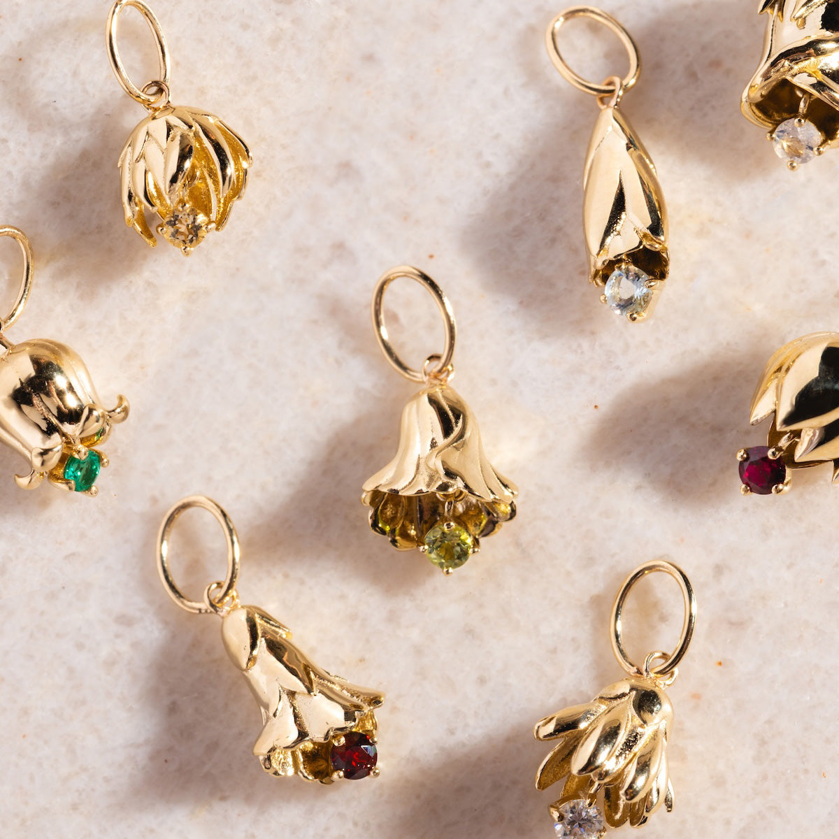 Solid Gold Birth Flower Bud Charms arranged on marble, each featuring a small colored gemstone to represent a birthstone. Ideal as a personalized pendant or a unique addition to your jewelry collection.