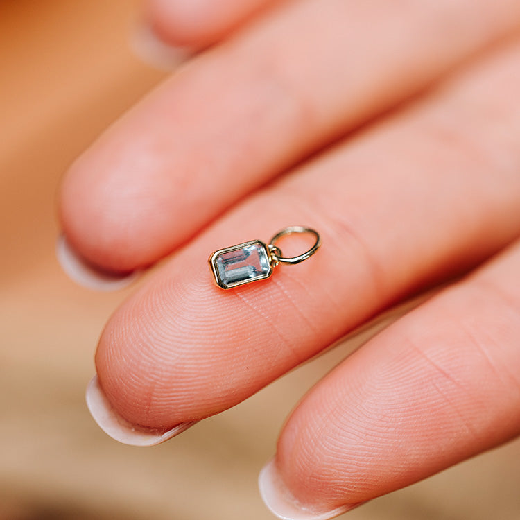 The Solid Gold Birthstone Baguette Charm, a small rectangular gemstone pendant with a gold frame and loop, rests on the tip of a persons neatly manicured finger.