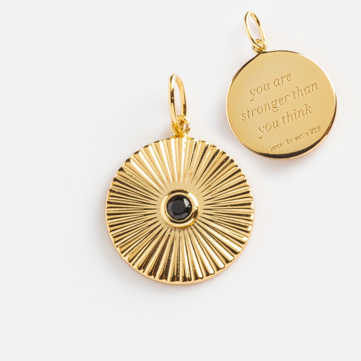 The Mantra Empowerment Coin Charm includes two round 14k gold plated pendants: one features a sunburst with a black center, the other is engraved you are stronger than you think, both with loops to attach to your chain.
