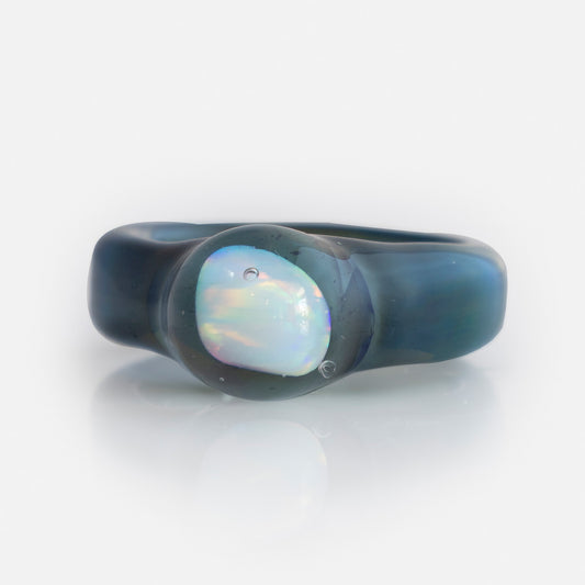 The Small Opal Glass Ring is a translucent dark blue borosilicate glass ring with a circular, iridescent opal-like stone set in the center, shown on a white background.