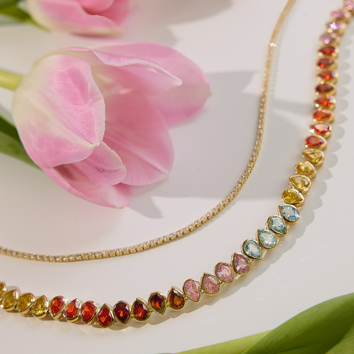 The Bold Bezel Set Tennis Necklace Multi Sample, featuring colorful oval gemstones in 14k yellow gold plating, is displayed on a white surface beside pink tulips next to a simple gold chain with sparkling cubic zirconia stones.