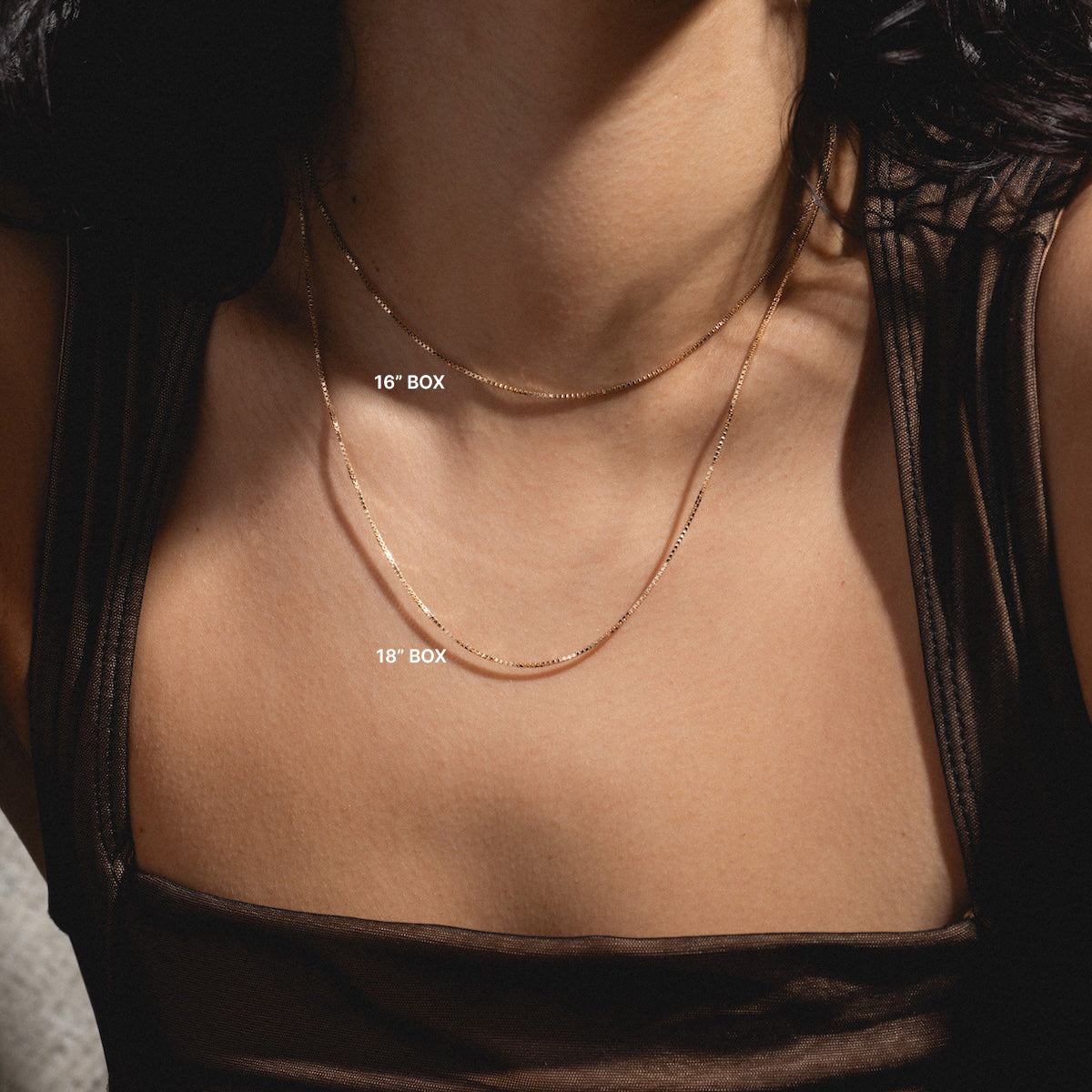 A person wearing a sheer dark top showcases two Solid Gold Chains for Charms, labeled “16” BOX” and “18” BOX,” elegantly layered on their collarbone and chest.