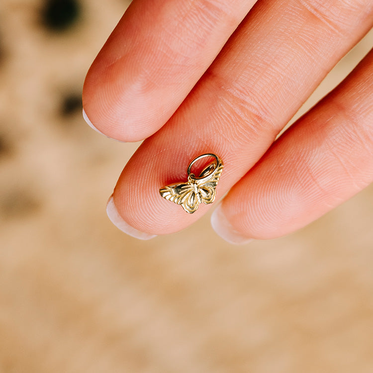 A hand with manicured nails holds the Solid Gold Nature Charm, shaped like a butterfly, between the thumb and index finger against a blurred beige background.