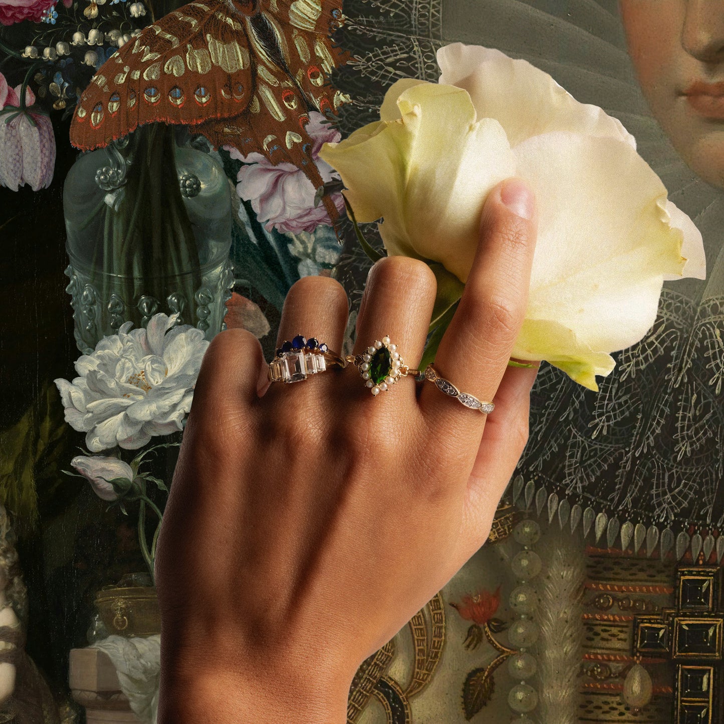 A hand wearing the 14k Blue Sapphire Arc Nesting Ring, along with two gemstone rings, holds a pale yellow rose before a richly detailed painting of flowers and a womans face in period dress.