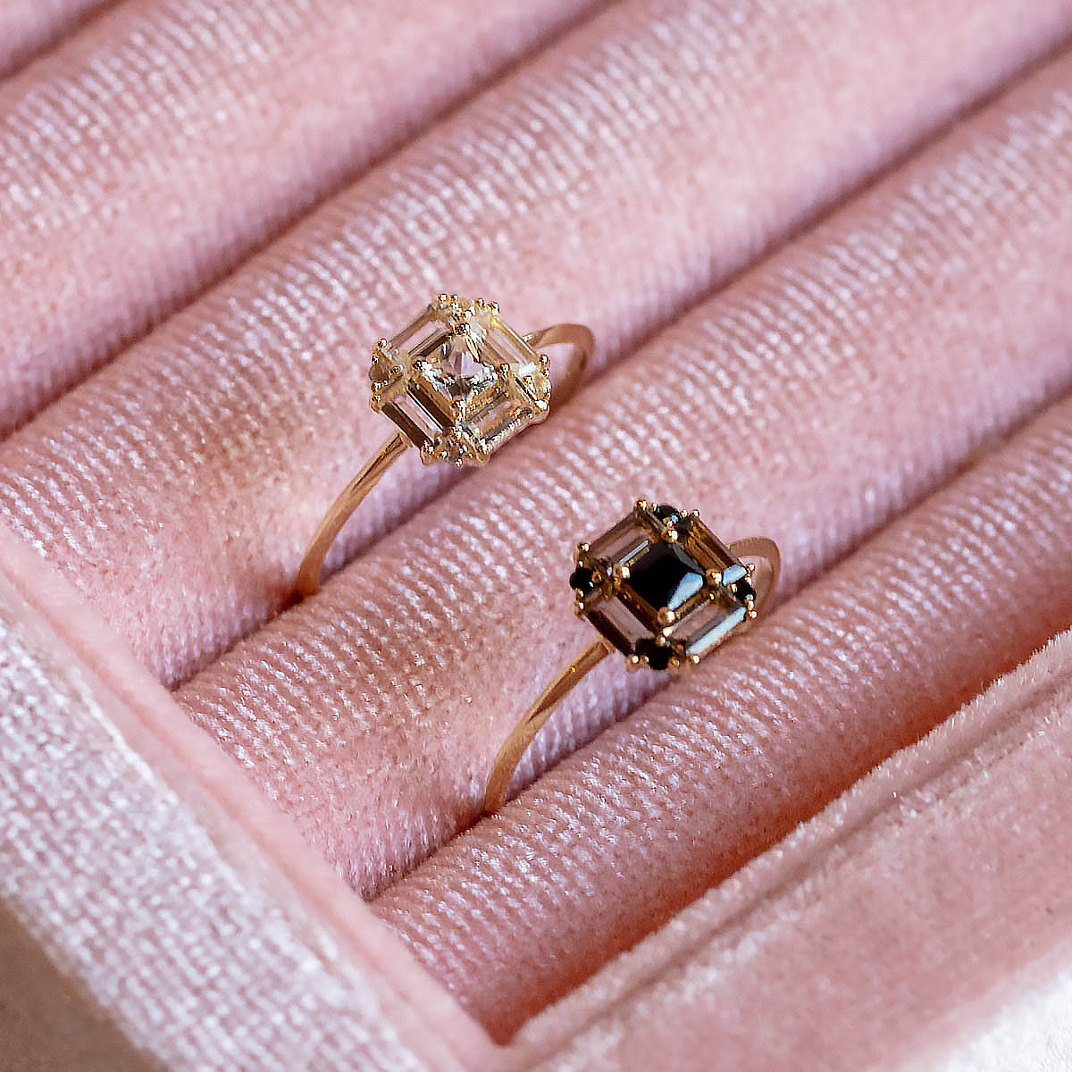 Two 14k gold rings from the Vintage Inspired Art Deco Engagement Ring collection are displayed in a pink velvet box; one features a clear gemstone and the other a dark stone, both set in geometric Art Deco designs.