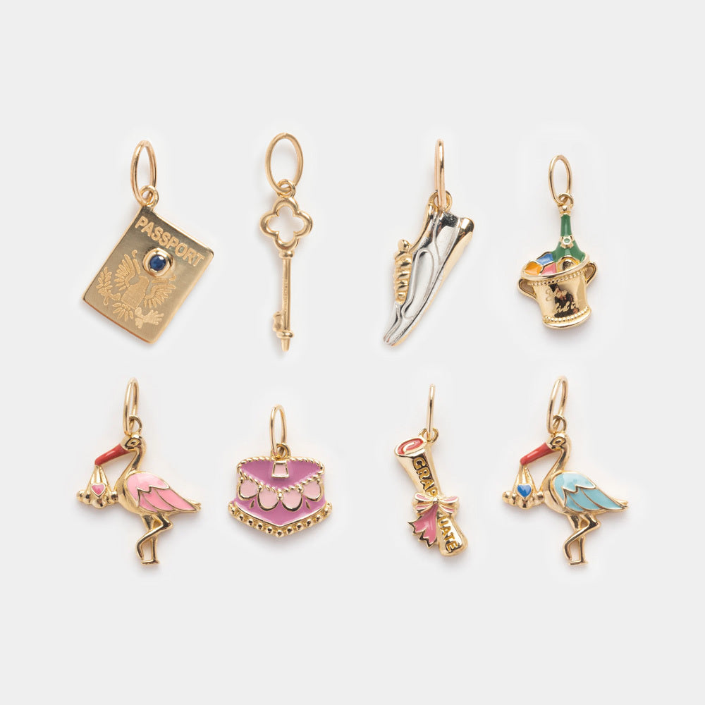 Eight Solid Gold Celebration Charms in 9k yellow gold, shaped like a passport, key, ballet slippers, champagne bucket, flamingo pair, purse, and engraved key with colorful enamel accents are displayed on a white background.