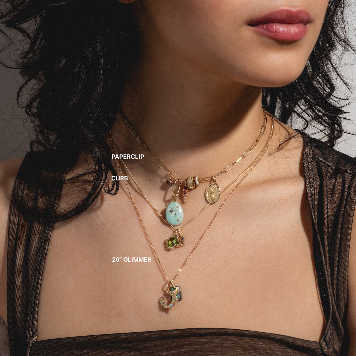 A woman wears a Solid Gold Chain for Charms, styled with multiple charms, a turquoise pendant on a curb chain, and a glimmering 20-inch chain with a rectangular green gemstone pendant.