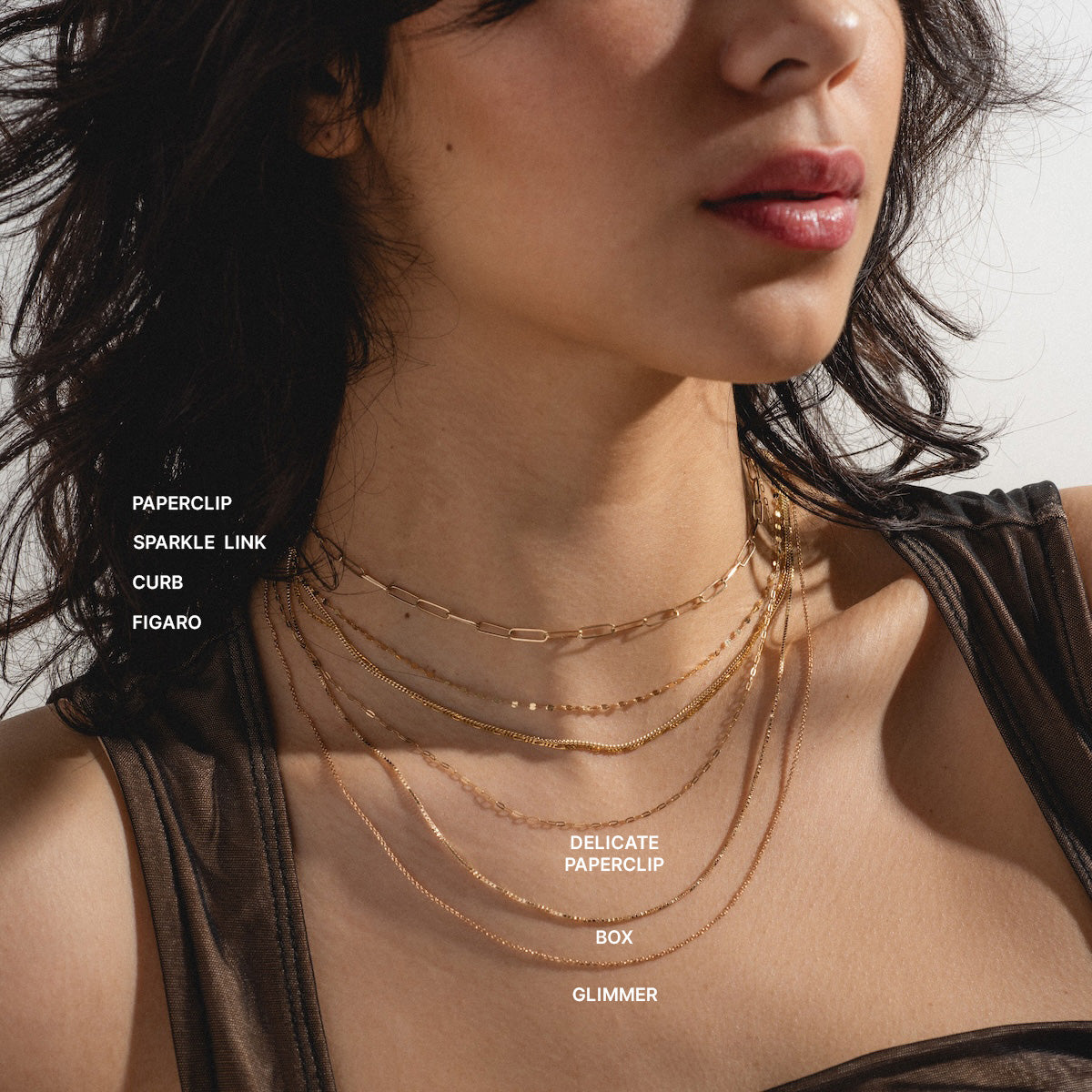 A woman with wavy dark hair in a sleeveless dark top wears the Solid Gold Chain for Charms, layered with other gold necklaces in styles like paperclip, sparkle link, curb, figaro, delicate paperclip, box, and glimmer.