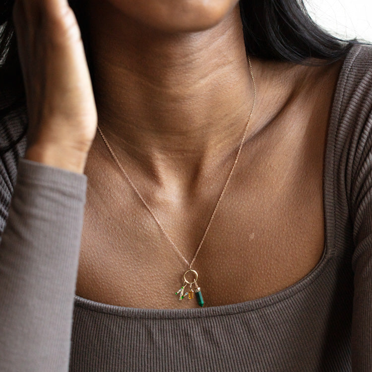 A woman wears a ribbed taupe long-sleeve top and showcases a Solid Gold Gemstone Letter Charm necklace adorned with several small, colorful charms. She touches her face, with her hair partially visible.