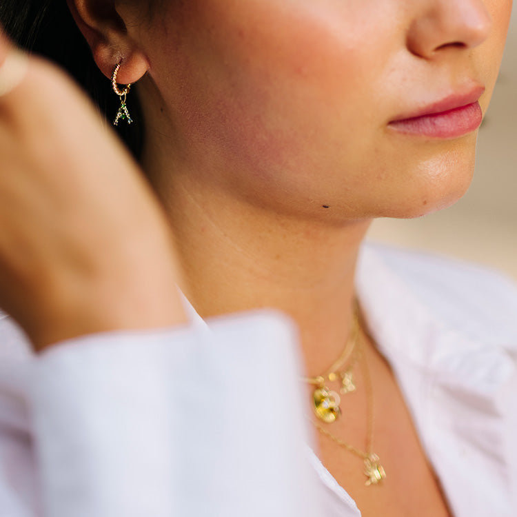 Close-up of a woman in a white shirt wearing gold hoop earrings with charms and layered necklaces, featuring the Solid Gold Gemstone Letter Charm. She touches her face, slightly out of focus, with softly pink lips.