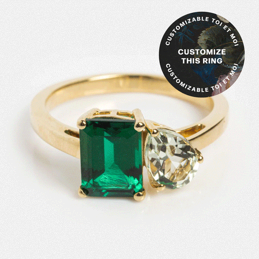 A solid gold Customizable Toi et Moi Ring with an emerald-cut green gemstone and a pear-shaped clear gemstone. A circular label reads: CUSTOMIZE THIS RING, customizable gemstone ring.