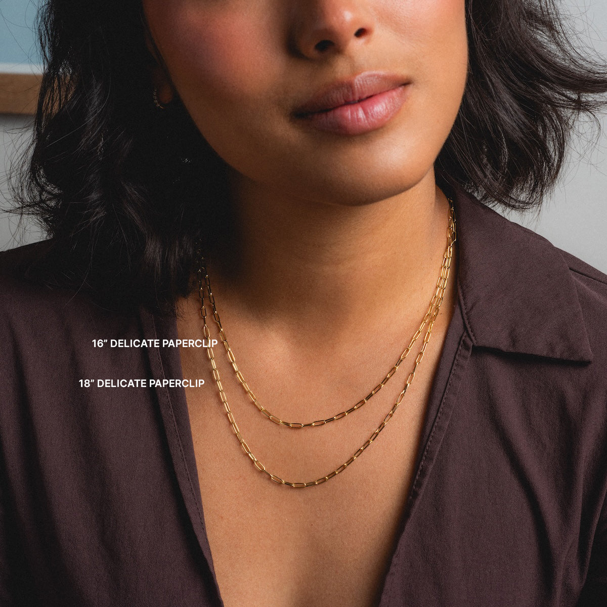 A woman in a dark brown shirt models two Chain For Charms gold paperclip chain necklaces—a 16 vermeil and an 18 delicate style—against a neutral background.