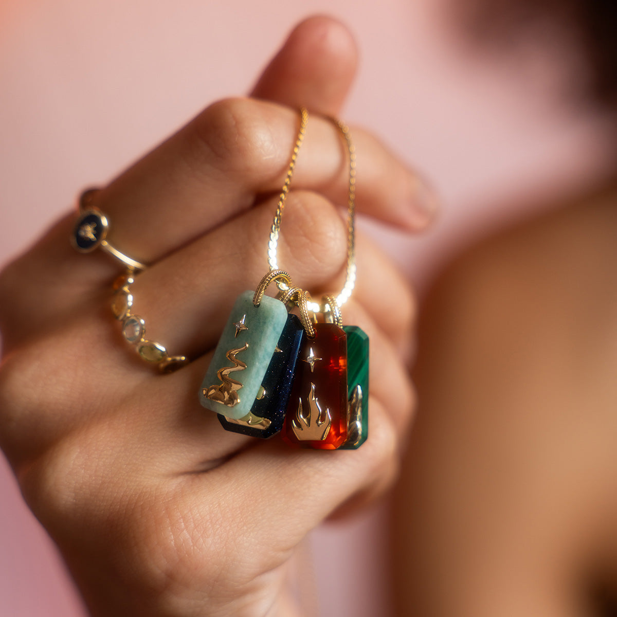 A hand holds three colorful gemstone pendants—a Solid Gold Crystal Element Charm and two natural crystal pendants—each on a gold chain with a unique gold symbol. The person also wears a gold ring with a blue stone and three clear stones.