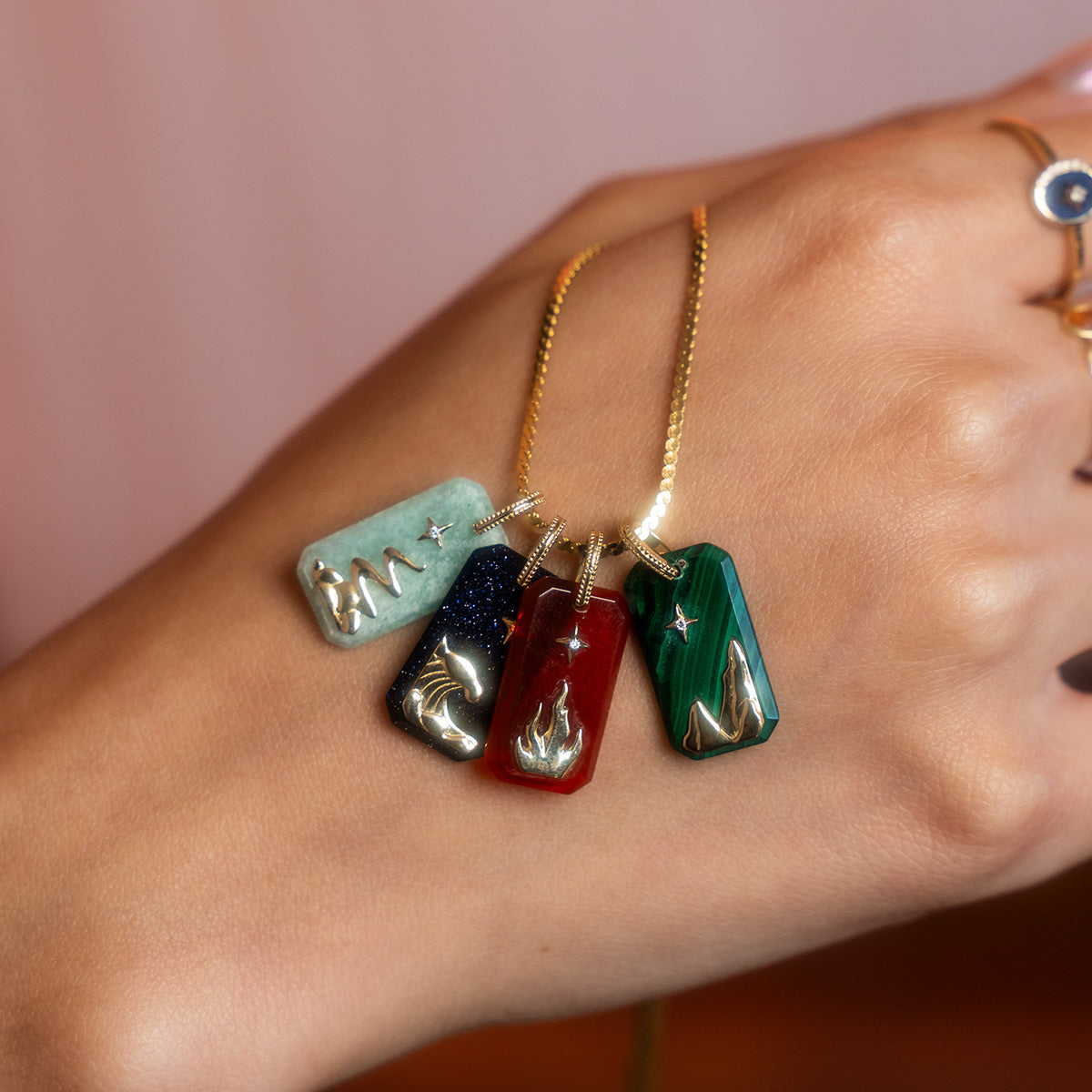 A hand displays four gold-chain necklaces with rectangular gemstone pendants—green, blue, red, and malachite—each featuring unique gold designs and Solid Gold Crystal Element Charm details. The hand is adorned with multiple gold rings.