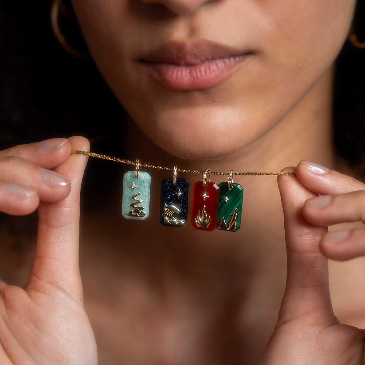 A person holds the Solid Gold Crystal Element Charm, a necklace with four vibrant rectangular gold pendants—a tree, wave, flame, and mountain—each symbolizing an element with colorful designs and metallic accents like a natural crystal pendant.