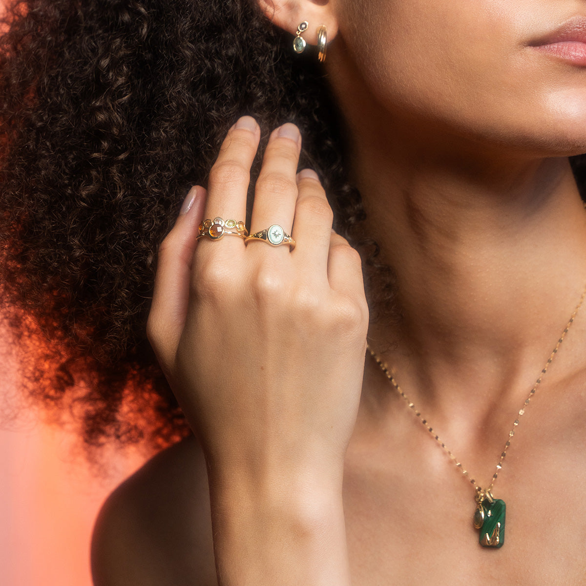 A woman with curly hair wears a gold necklace featuring the Solid Gold Crystal Element Charm. She also has on gemstone rings and small gold hoops, raising her hand near her face to display the jewelry details.