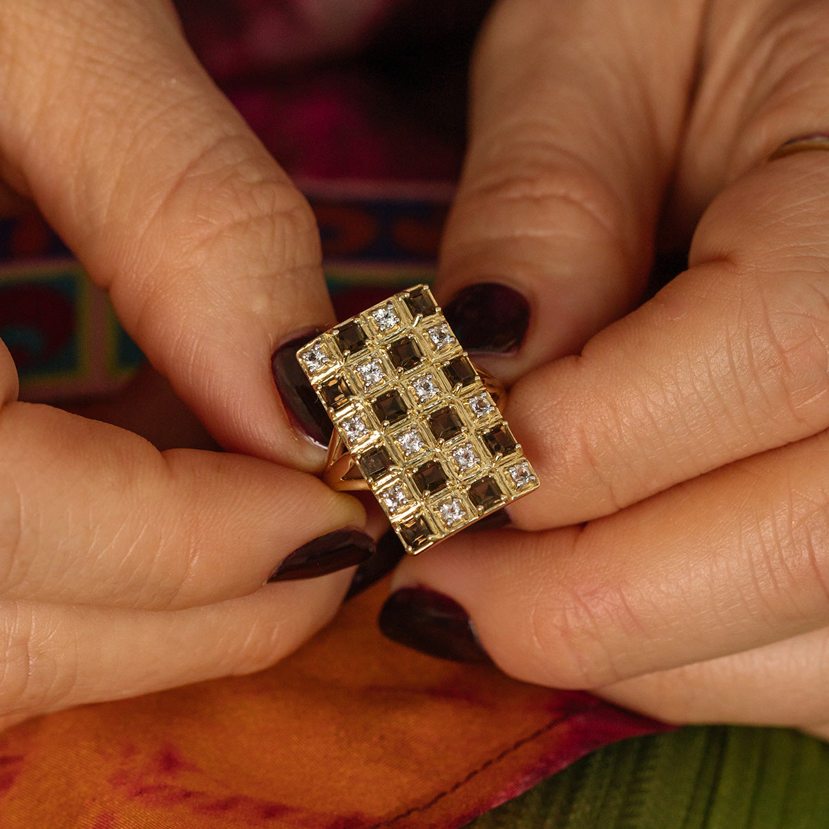 A person with dark nail polish holds the Solid Gold Checkerboard Gemstone Shield Ring (sample size 7), featuring a large rectangular 9k yellow gold band set with sparkling white topaz gems, against colorful fabric.