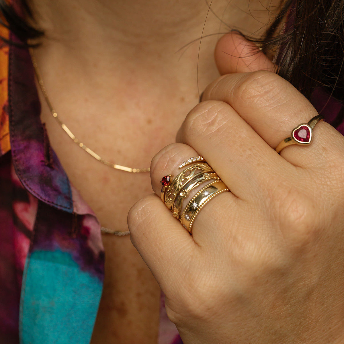 A close-up of a hand wearing the Solid Gold Bezel Set Ruby Heart Ring and a gold band, paired with a gold necklace. The person is dressed in a colorful, patterned shirt.