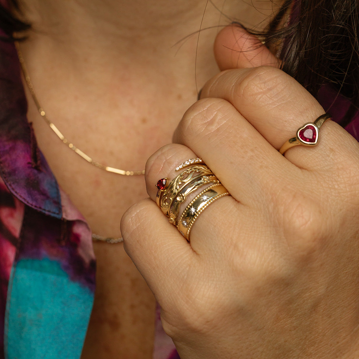 A close-up of a hand adorned with multiple gold rings, including the Solid Gold Mixed Metal Filigree Band, plus a gold necklace and a colorful patterned shirt.
