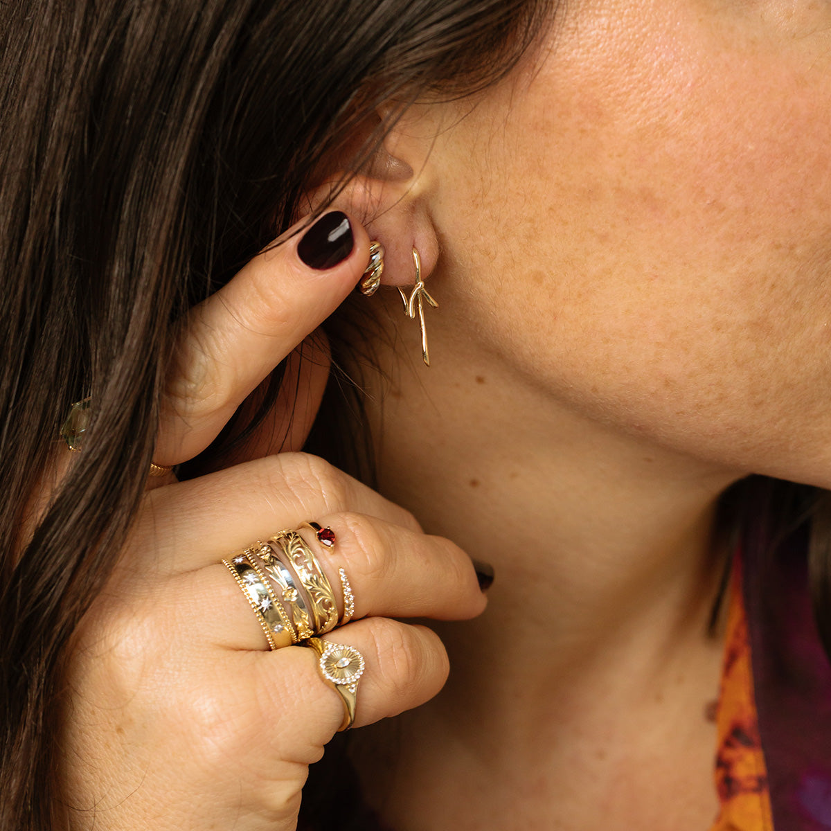 A close-up of a womans hand wearing multiple gold rings—including the Solid Gold White Topaz Etched Signet Pinky Ring—shows her dark nails, gold earrings, and brown hair.